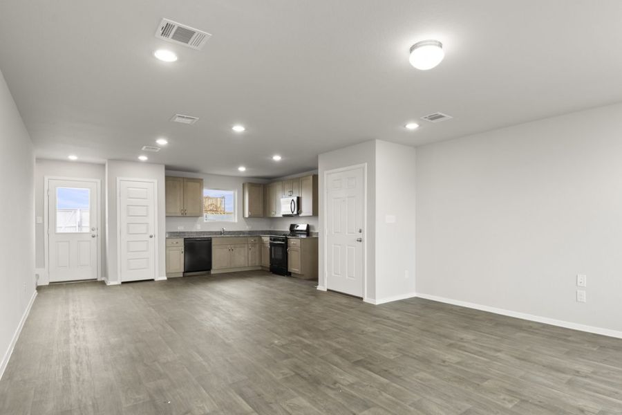 Image of a cottage home living room with brown vinyl flooring, light grey painted walls and a L-shaped kitchen in the distance
