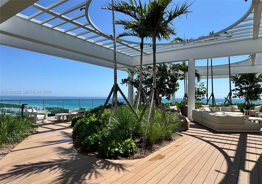 Expansive outdoor terrace featuring wood-finish decking, a white slatted pergola, lush landscaping, and a clear glass railing