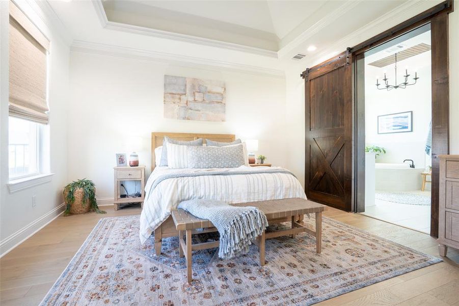 Bedroom featuring light wood finished floors, a barn door, ornamental molding, a tray ceiling, and hanging lights Bedroom featuring light wood finished floors, a barn door, ornamental molding, a tray ceiling, and hanging lights