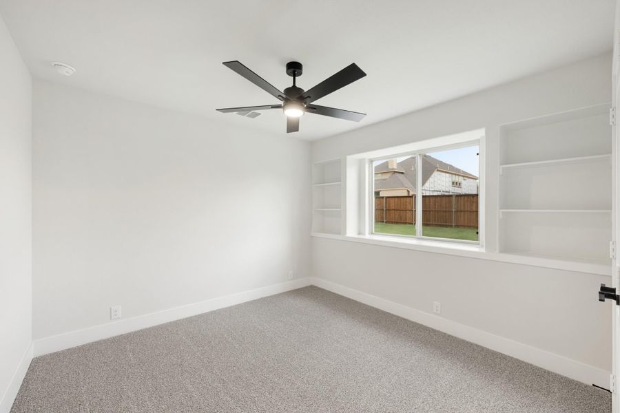 Empty bedroom with gray carpet, black ceiling fan, built-in shelves, and a window overlooking a fenced backyard