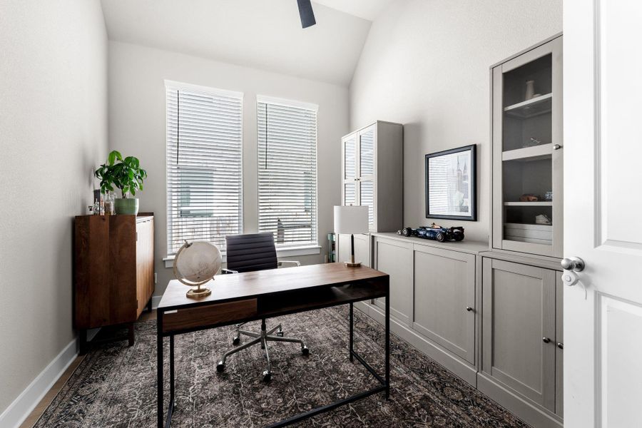Office space featuring dual windows with blinds, built-in cabinetry with glass-front display sections, and neutral wall tones