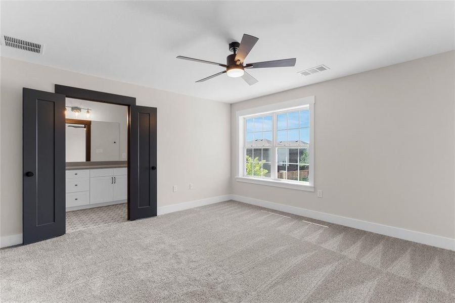 Carpeted room featuring a ceiling fan with integrated lighting, a large window with white trim, and dark double doors opening to a bathroom with a white vanity and gray countertop