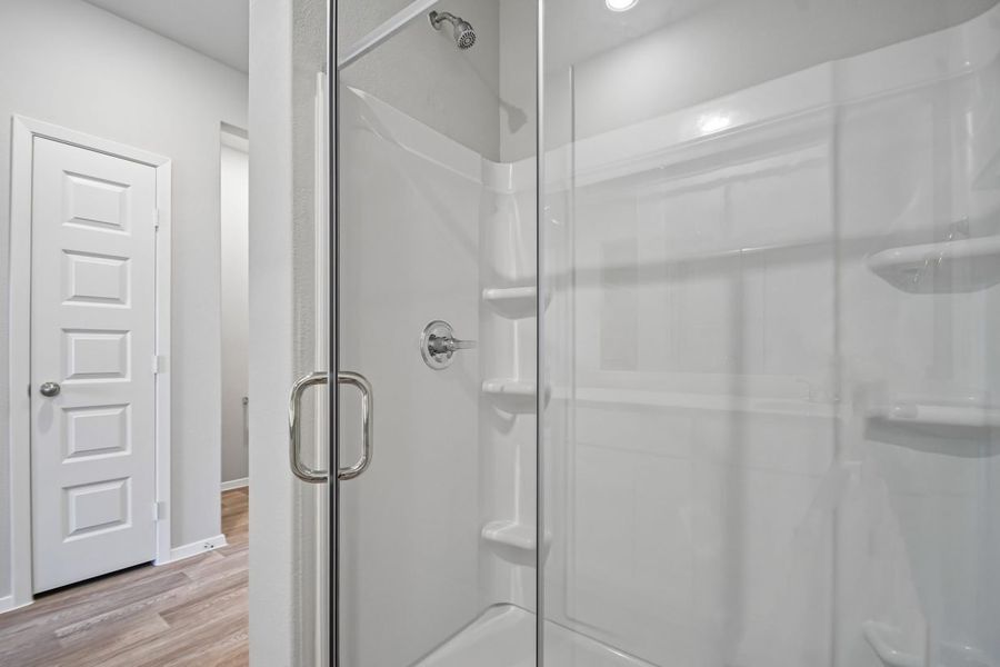 Image of walk-in shower with white surround and glass in front with chrome fixtures. Small white door to linen closet and a peek into the water closet. White walls and wood-look flooring