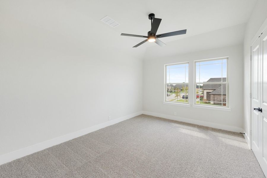 Empty bedroom with carpet flooring, black ceiling fan, and two windows overlooking a neighborhood