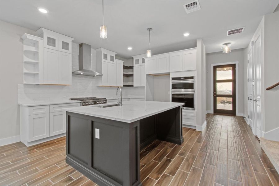 Kitchen featuring a center island with sink, tasteful backsplash, wall chimney exhaust hood, double oven, and white cabinets