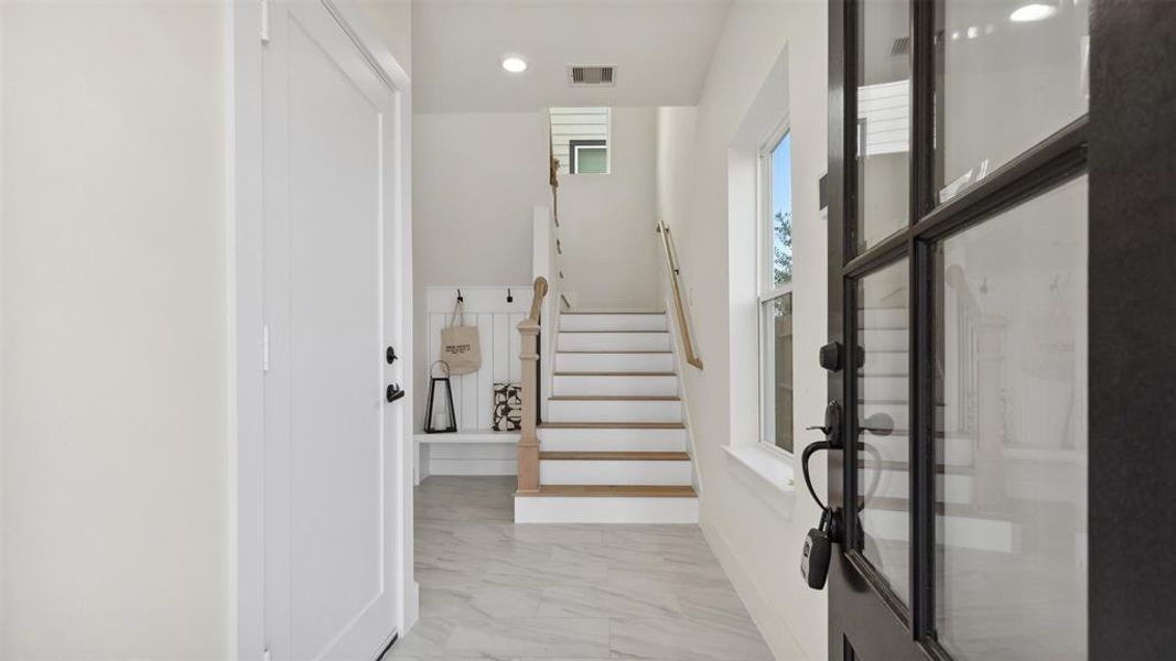 Enter thru the front door to be greeted by bright, clean entryway with a staircase leading up. The space features modern finishes with a tiled floor, natural light from a window, and a welcoming, neutral color palette.