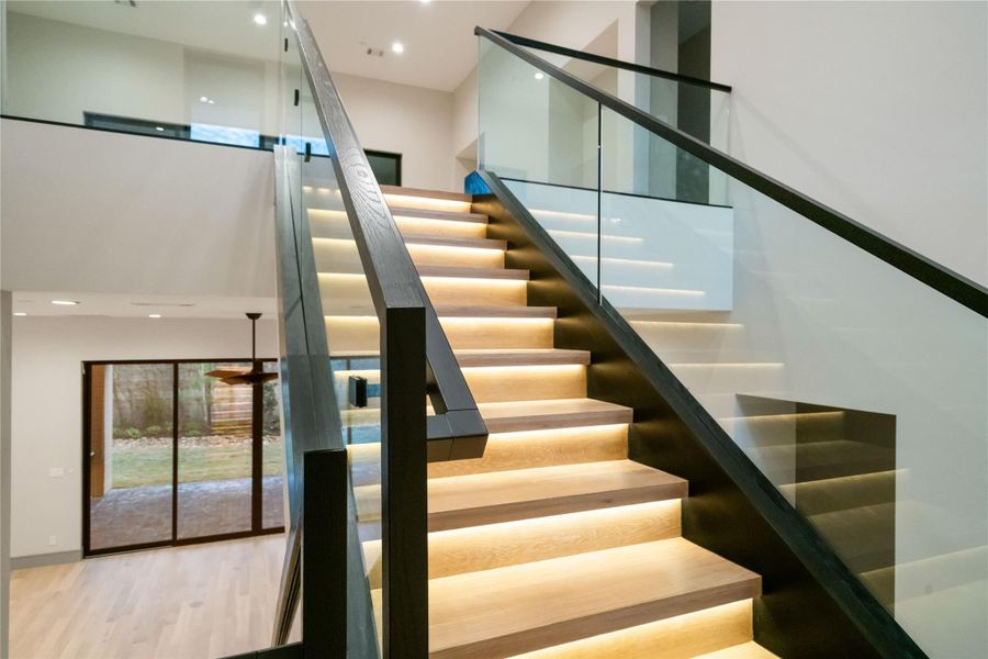 This photo showcases a modern staircase with sleek glass railings and elegant under-step lighting. The open layout includes a view of a spacious room with sliding glass doors leading to an outdoor space, highlighting natural light and contemporary design.