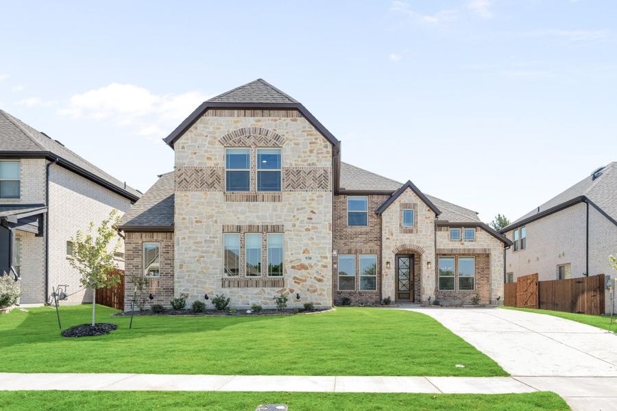 Two-story home with stone and brick exterior, dark roof, and green front lawn with concrete driveway