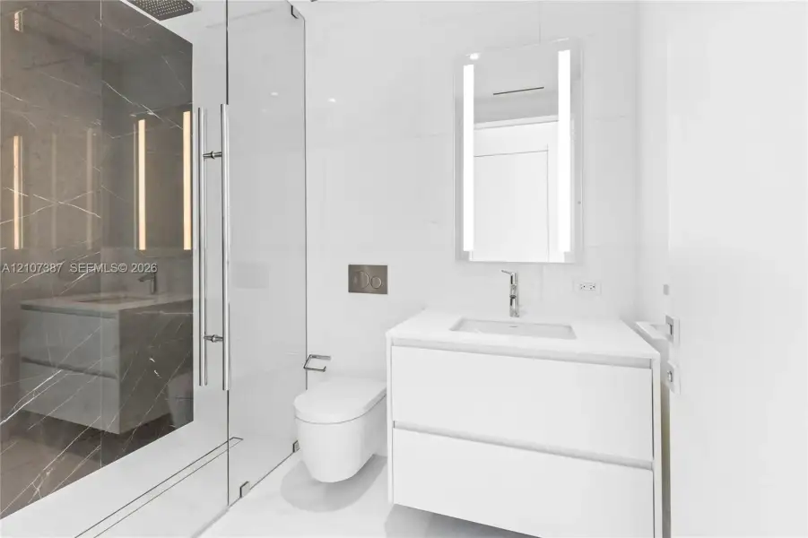 Floating white vanity with solid surface countertop, wall-mounted toilet, glass-enclosed shower featuring dark stone-finish tile, and integrated LED-lit vanity mirror