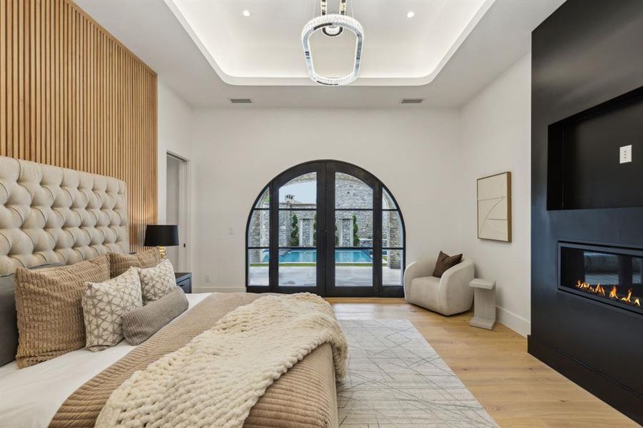 Bedroom with access to courtyard through iron and glass French doors , designer lighting, a tray ceiling, white oak wood finished floors, a linear fireplace and wood-paneled accent wall Bedroom with access to courtyard through iron and glass French doors , designer lighting, a tray ceiling, white oak wood finished floors, a linear fireplace and wood-paneled accent wall