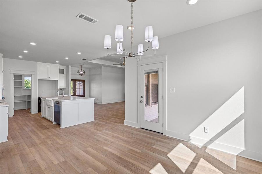 Open-concept living space featuring wood-finish flooring, a contemporary chandelier, a kitchen island with an integrated sink, white cabinetry, and a pantry with built-in shelving