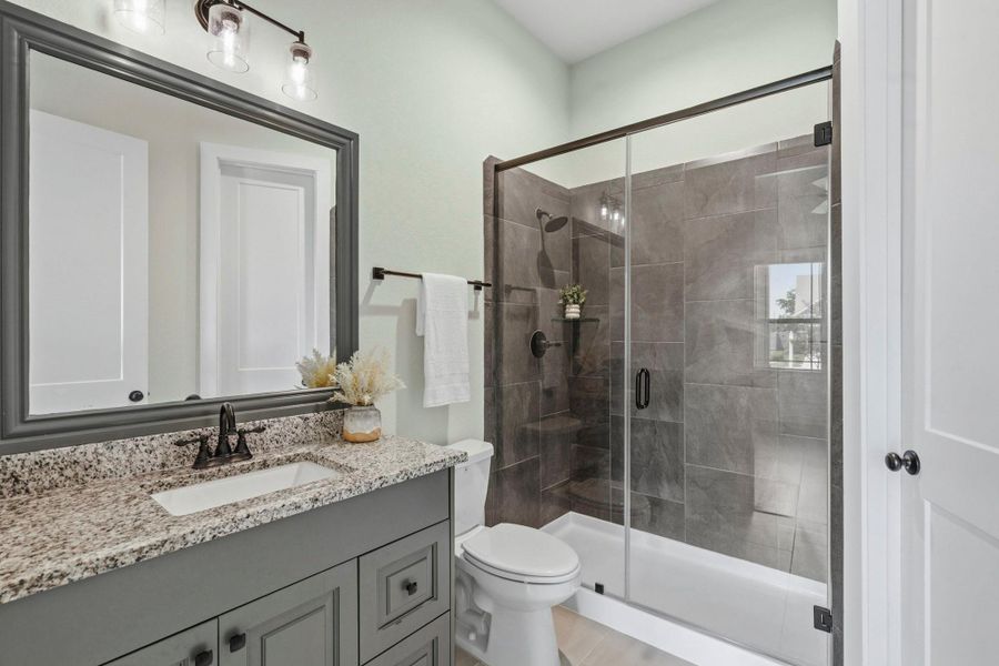 This beautifully appointed Secondary Ensuite Bath combines timeless elegance with everyday functionality. Featuring an undermount sink, sleek granite countertops, and a framed mirror, the space exudes a refined and polished aesthetic. The walk-in shower serves as a striking focal point, enveloped in rich, dark tile that creates a sophisticated, spa-like atmosphere and adds a sense of depth and luxury to the room.