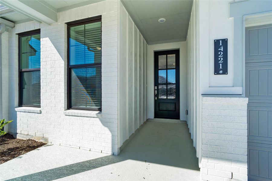 This covered front porch exudes modern curb appeal, showcasing crisp white brick and a stately black-framed glass front door.