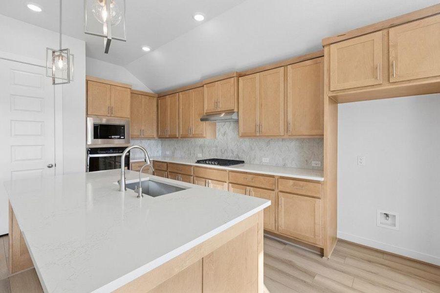 Kitchen featuring light brown cabinets, vaulted ceiling, decorative light fixtures, a center island with sink, and backsplash