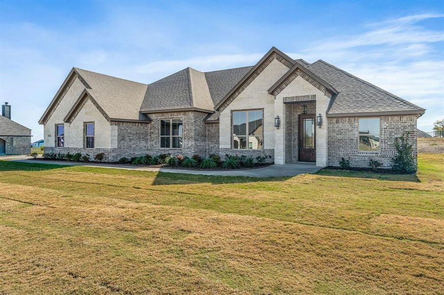 French country style house with stone siding, a front yard, roof with shingles, and brick siding
