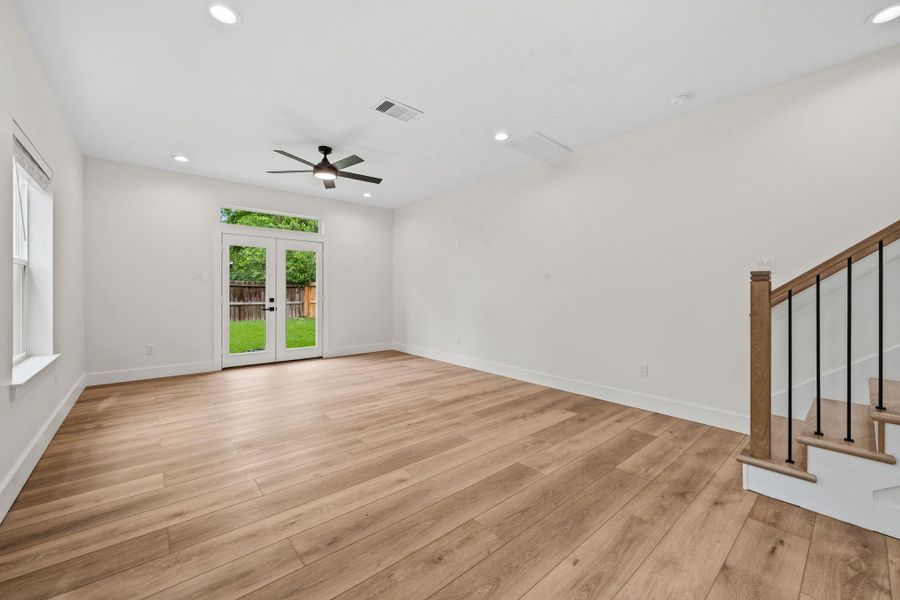 Spacious living area with elegant wood flooring, featuring a modern ceiling fan and abundant natural light from large windows and French doors leading to a lush backyard.