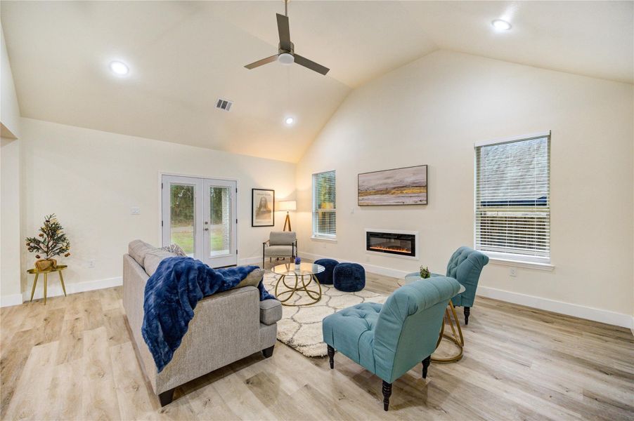 This spacious living room features vaulted ceilings, a modern ceiling fan, and plenty of natural light. It includes a cozy electric fireplace, stylish seating, and French doors leading to the outdoors, all set on elegant wood flooring.
