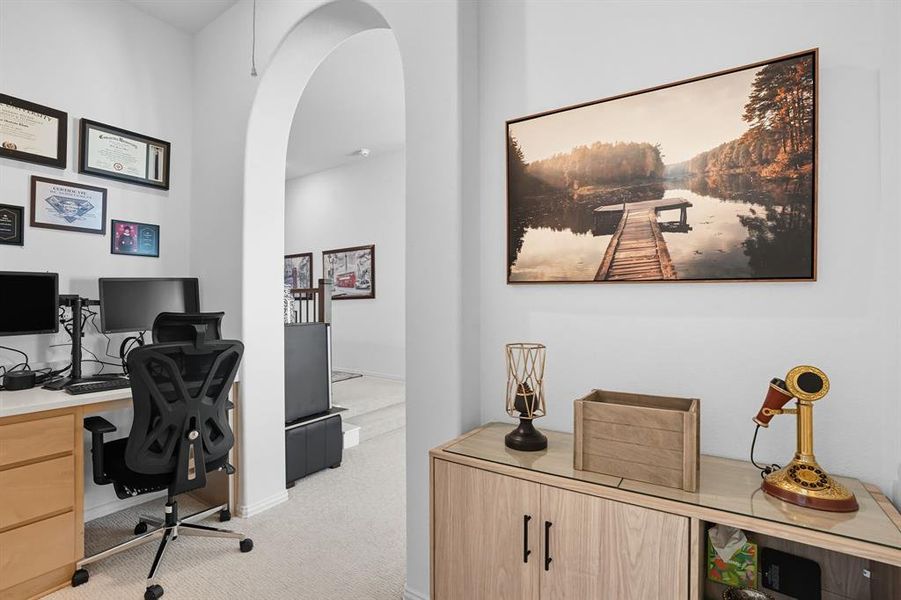 Carpeted office area featuring a light wood-finish desk with drawers, an arched doorway, and built-in cabinetry with wood-finish doors and dark hardware