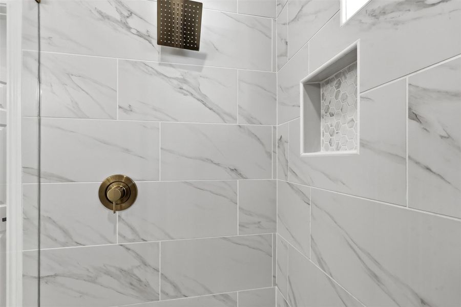 Primary bathroom shower detail showcasing white marble tile walls, built in niche, and gold fixtures.