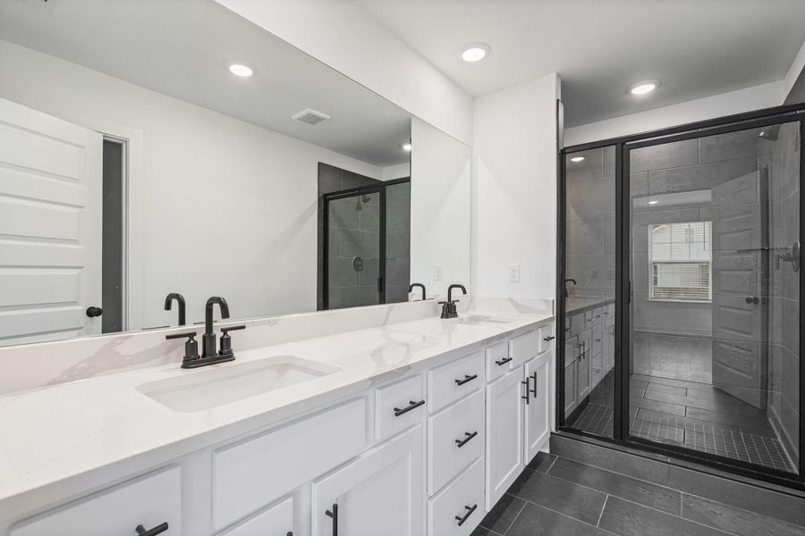 Dual vanity with integrated sinks and black matte fixtures, white cabinetry, a large frameless mirror, and a glass-enclosed shower with dark-toned tile Dual vanity with integrated sinks and black matte fixtures, white cabinetry, a large frameless mirror, and a glass-enclosed shower with dark-toned tile