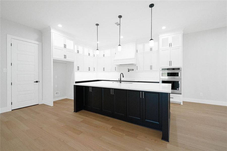 Kitchen featuring dual tone cabinets, a kitchen island with sink, light wood-style floors, double oven, and hanging light fixtures