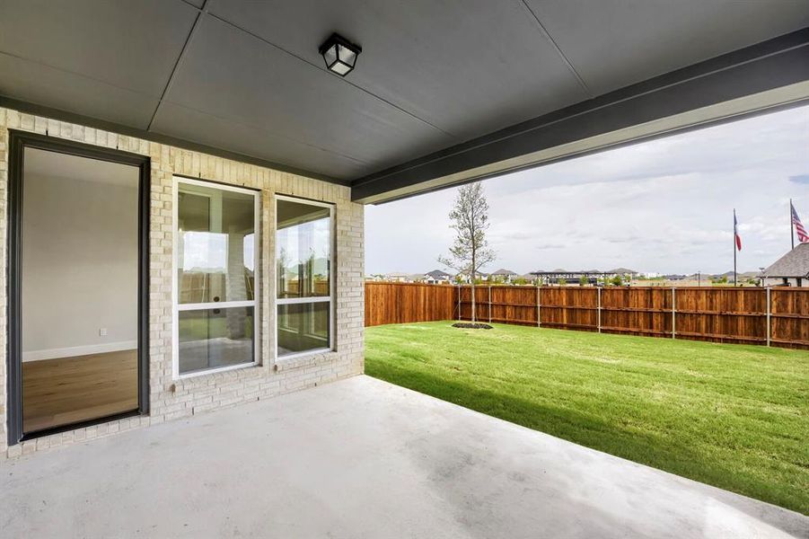 Covered concrete patio with light brick exterior and a fenced backyard featuring a young tree and manicured lawn
