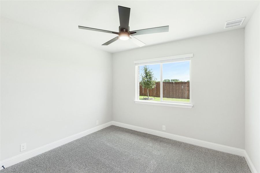 Empty bedroom with gray carpet, ceiling fan, and window overlooking fenced backyard