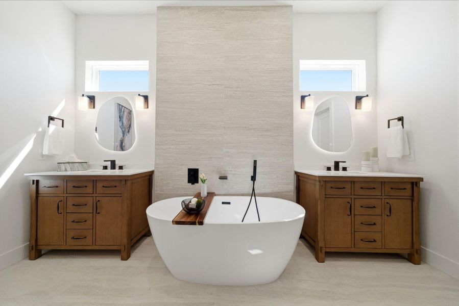 This bathroom features a modern design with a freestanding tub, dual wooden vanities, and stylish oval mirrors. It offers abundant natural light from two small windows and is enhanced by elegant fixtures and a neutral color palette.