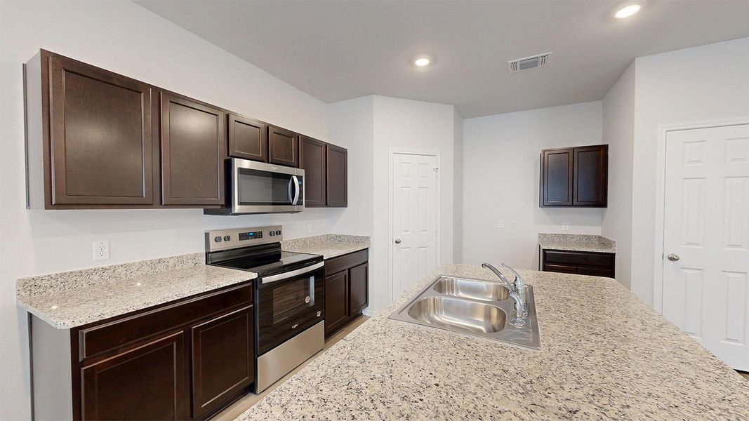 Kitchen featuring stainless steel appliances, dark wood finish cabinetry, light stone counters, recessed lighting, and an island with sink