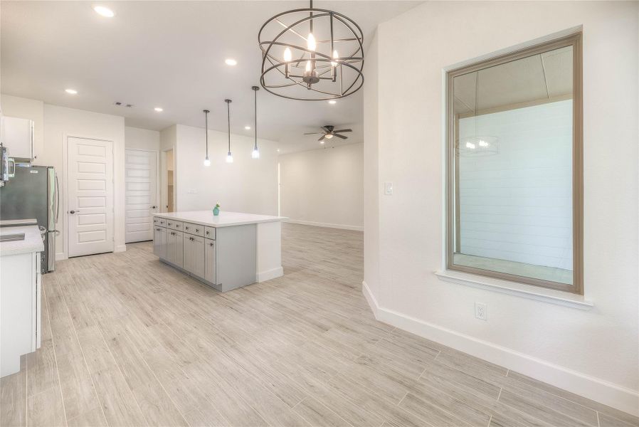 This spacious, modern kitchen features light wood flooring, a large island with storage, stainless steel appliances, and pendant lighting. An open floor plan flows into a bright living area with a ceiling fan, perfect for entertaining.