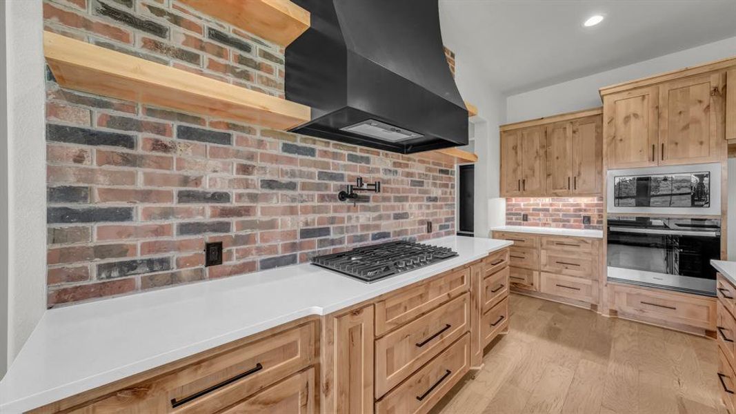 Kitchen featuring a gas range cooktop, brick backsplash, wood-finish flooring, light wood cabinetry, and integrated stainless steel appliances