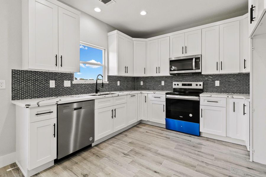 This modern kitchen features sleek white cabinetry with black hardware, a stylish gray tile backsplash, and stainless steel appliances, including a dishwasher and oven. The bright, airy space is enhanced by natural light from a window, complemented by light wood flooring for a contemporary feel.