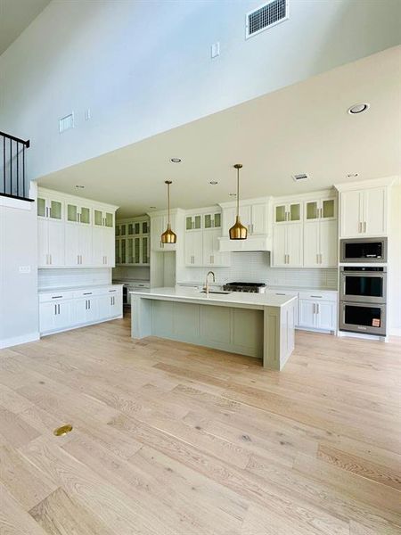 Kitchen featuring glass insert cabinets, white cabinetry, an island with sink, and light wood-style floors Kitchen featuring glass insert cabinets, white cabinetry, an island with sink, and light wood-style floors