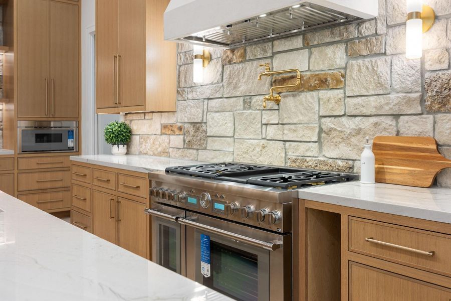 A close-up perspective reveals a thoughtfully designed cooking space where texture and detail take center stage. The natural stone backsplash adds depth and character, paired with warm wood cabinetry for a timeless look. The highlight is the integrated pot filler, adding both convenience and elevated design, while the clean lines and layered materials create a polished and functional culinary environment.