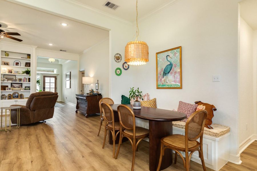 This inviting space features a cozy dining nook with a built-in bench and stylish pendant light, leading into a living area with custom built-in shelves and a comfortable seating arrangement. The open layout and light wood look flooring create a warm, welcoming atmosphere.