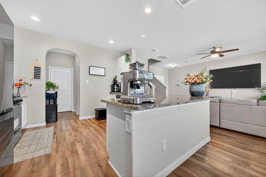 This photo showcases a modern, open-concept living area with hardwood floors, neutral walls, and recessed lighting. The space features a kitchen island with a coffee machine, a cozy seating area with a ceiling fan, and decorative plants, creating a welcoming atmosphere.