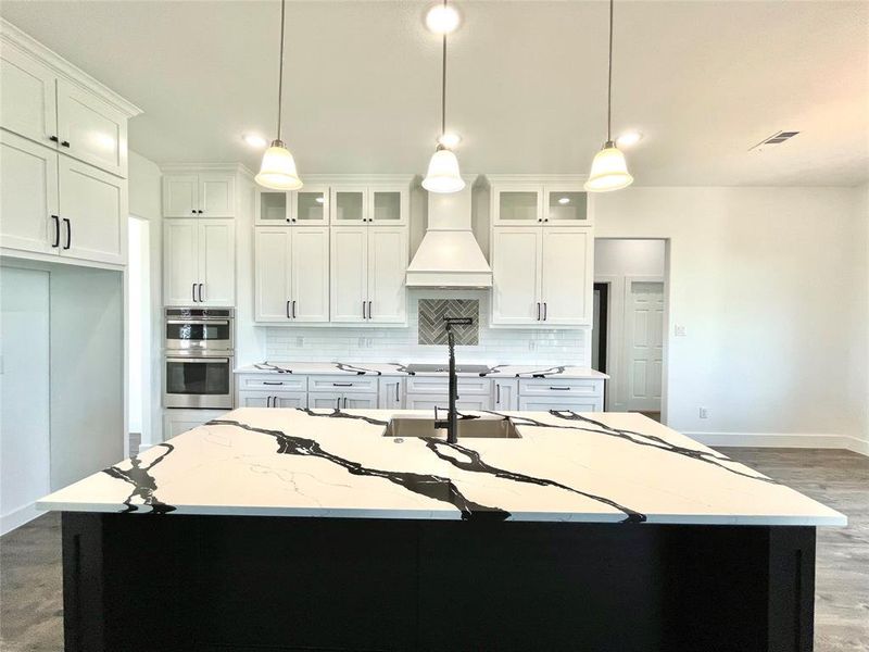 Kitchen with a kitchen island with sink, baseboards, premium range hood, backsplash, and double oven Kitchen with a kitchen island with sink, baseboards, premium range hood, backsplash, and double oven