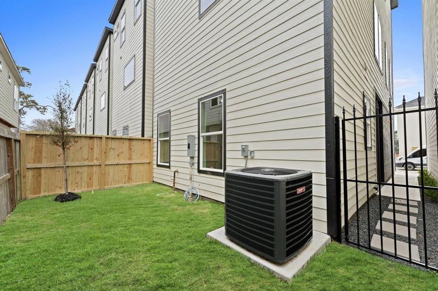 This photo shows a narrow side yard with well-maintained grass, a wooden privacy fence, and a small tree. The exterior of the house is visible with light-colored siding, and there's an air conditioning unit on a concrete pad. A black metal gate leads to a pathway along the side of the house.