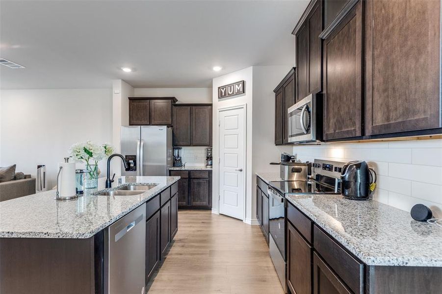 Kitchen featuring dark brown cabinets, appliances with stainless steel finishes, decorative backsplash, an island with sink, and light stone counters Kitchen featuring dark brown cabinets, appliances with stainless steel finishes, decorative backsplash, an island with sink, and light stone counters