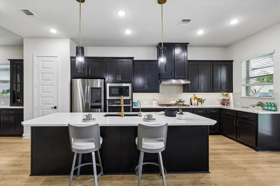Modern kitchen featuring a large central island with seating, dark cabinetry, stainless steel appliances, light countertops, and wood-finish flooring Modern kitchen featuring a large central island with seating, dark cabinetry, stainless steel appliances, light countertops, and wood-finish flooring