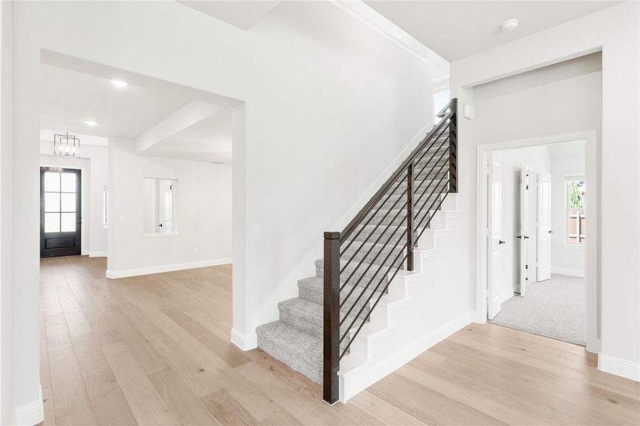 Open foyer with carpeted staircase, dark metal railing, light hardwood floors, and black front door visible in background.