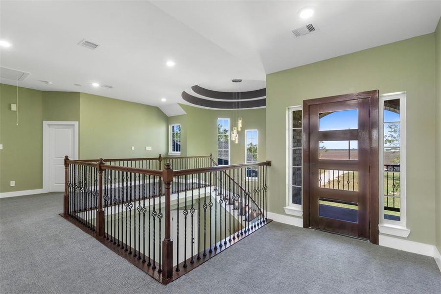 This photo showcases a spacious, well-lit upstairs area with a modern open railing design and a stylish front door. The room features soft green walls, recessed lighting, and carpeted flooring, creating a welcoming atmosphere. Multiple windows allow for plenty of natural light.