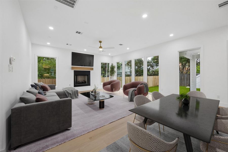 The main living area was designed to feel connected to both the interior and outdoor spaces of the home, with oversized rear-facing windows bringing in views of the surrounding greenery and backyard beyond. Clean architectural lines, wide-plank engineered wood flooring, and the open scale of the room create a setting that feels bright, comfortable, and well suited for both everyday living and larger gatherings. (digitally staged)