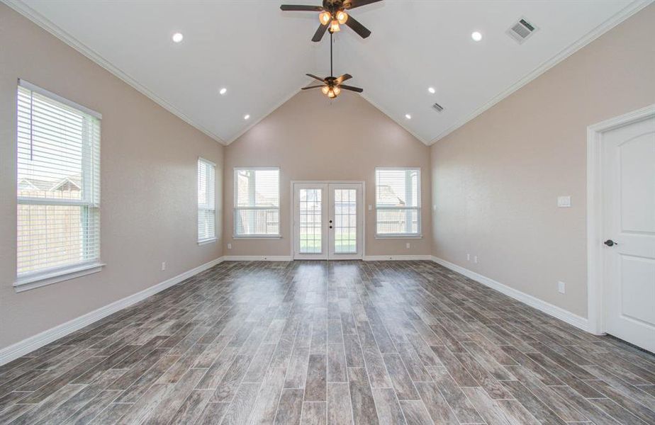 This is a spacious and well-lit room featuring wood-look tile flooring, a high vaulted ceiling with recessed lighting and a ceiling fan, multiple windows for natural light, and a glass door leading to the outside.