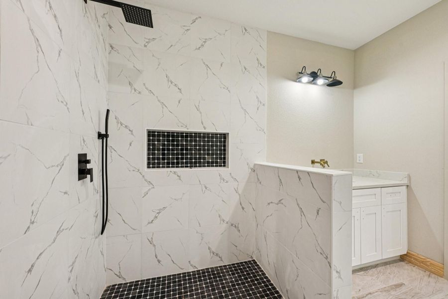 This photo showcases the  open shower area featuring patterned tiles and a black rainfall shower-head. There is a built-in black tile niche for storage.