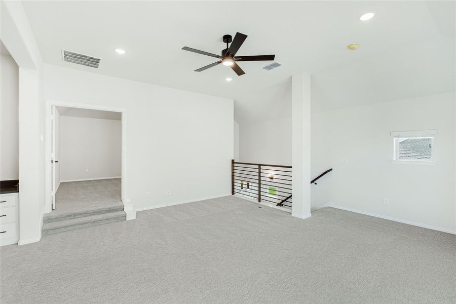 Open upstairs loft area with gray carpet, ceiling fan, and metal stair railing overlooking lower level