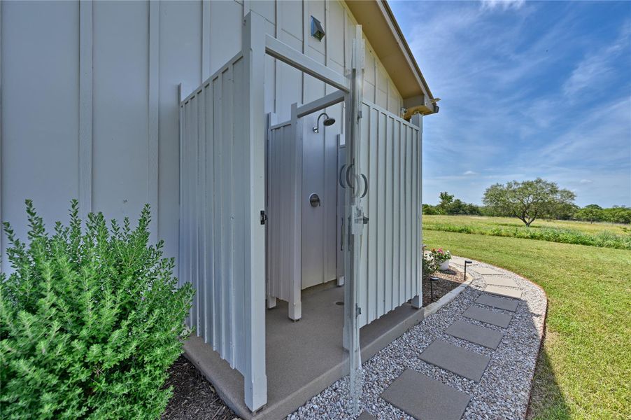 An outdoor shower and changing area awaits, nestled beside a gravel and stepping stone path leading to a verdant field.