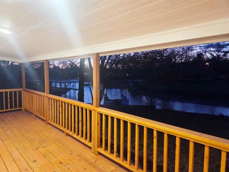 Expansive covered deck featuring natural wood flooring and railings, overhead recessed lighting, and a tranquil water feature