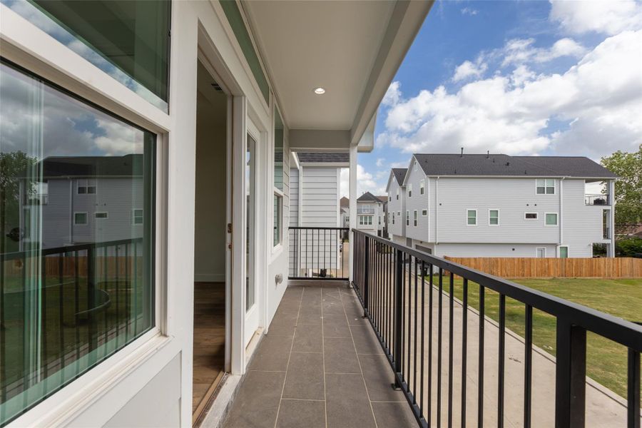 This photo features a modern balcony with sleek railing, overlooking a suburban neighborhood. The space connects to the interior through a glass door, offering a view of clear skies. Ideal for relaxation and enjoying outdoor views.