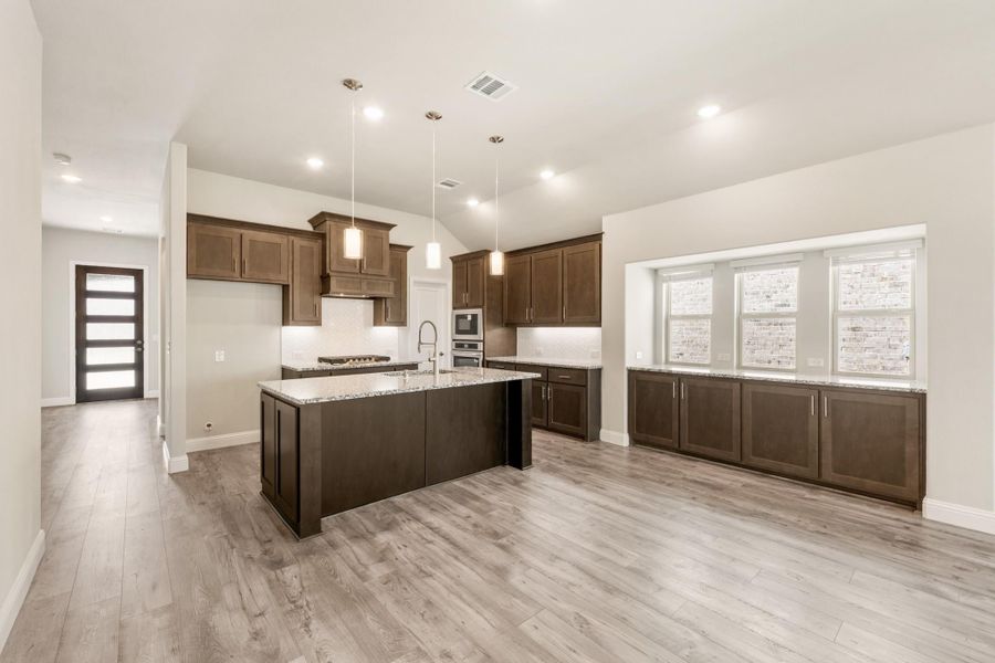 Kitchen with dark brown cabinets, granite island, pendant lights, and light wood floors